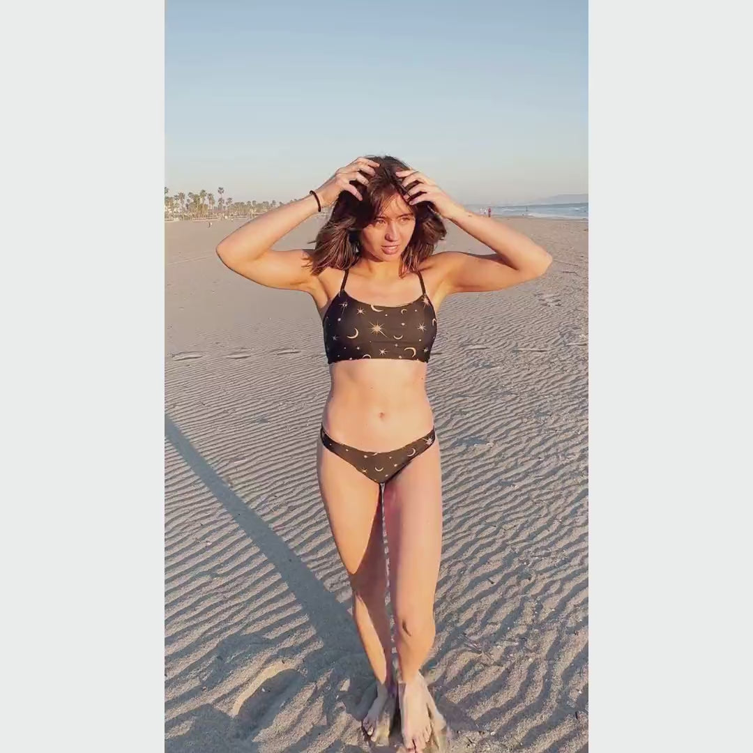 Woman on beach in Santa Monica wearing a gold, lilac and black cosmic stars and moon print bikini with black straps 