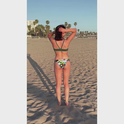 Athletic woman on beach in Santa Monica doing a twirl wearing a sport bikini with a green, lilac, yellow, orange and black tropical flower print and dark green straps.