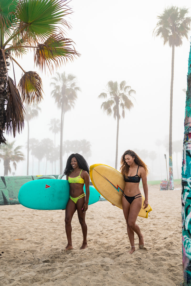 Two girls walking on Venice beach near graffiti walls with surfboards smiling wearing Pitviper bikinis 
