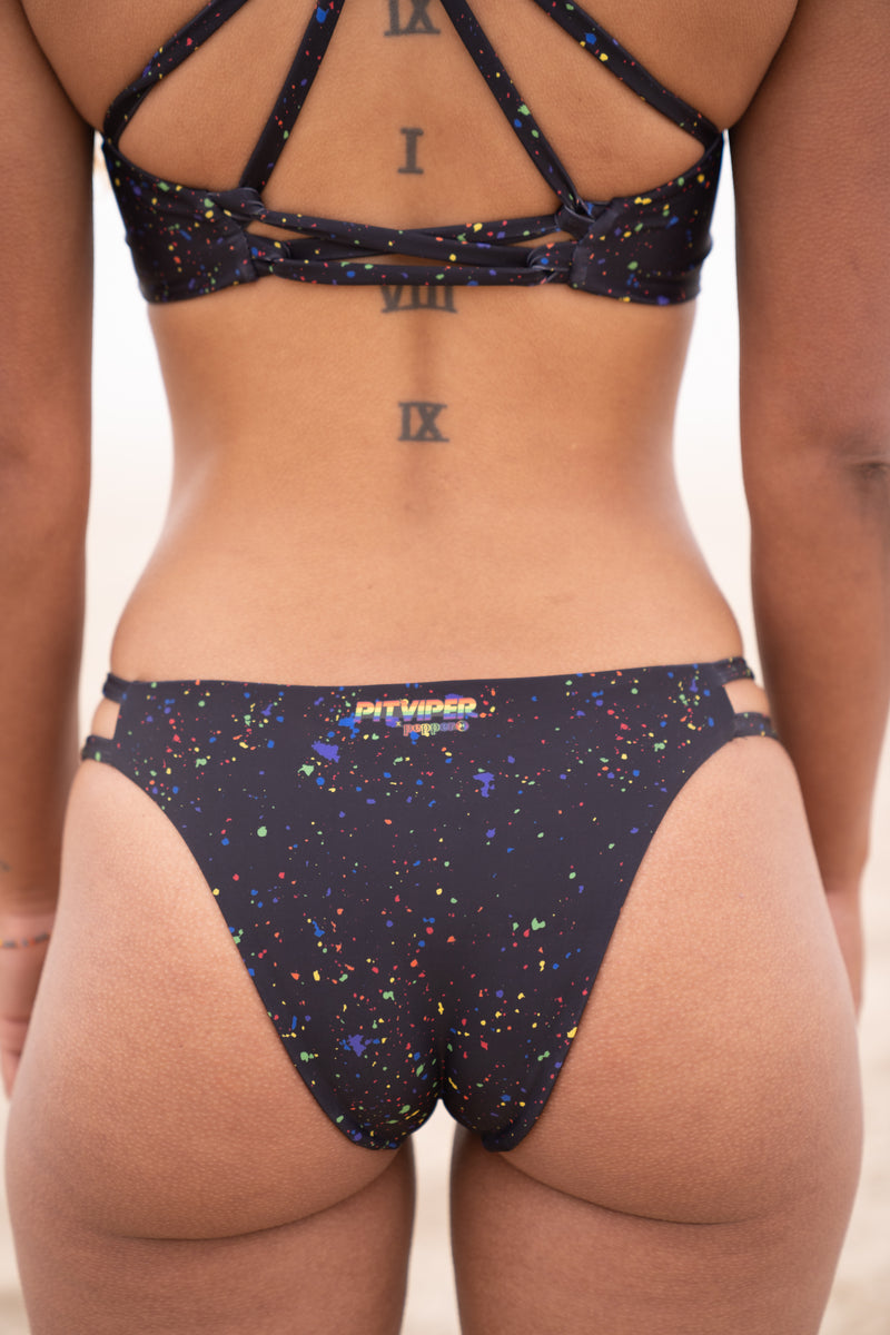 Backside of woman wearing Pitviper Rollerdome bikini with black and rainbow splatter print. Criss cross star shaped back design, cheeky but not too cheeky bottoms