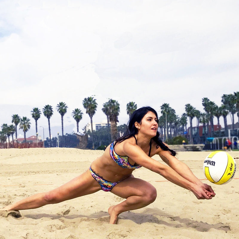 A female beach volleyball player dives for a volleyball wearing a colorful geometric print sport bikini with black straps.
